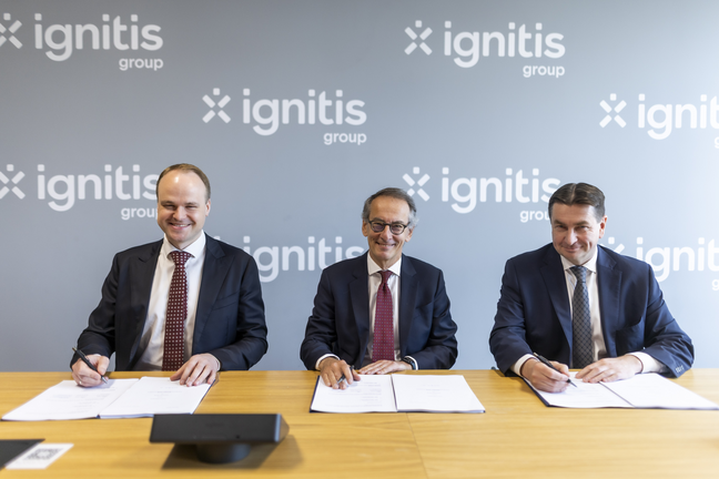 Trys kostiumuoti vyrai prie stalo, visi šypsosi ir pasirašo dokumentus, fonas su „Ignitis Group“ logotipu.
