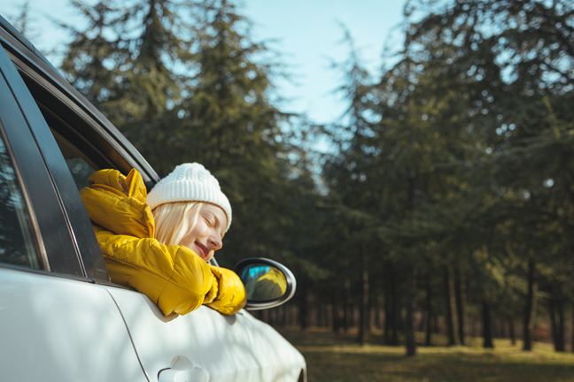 Mežā stāv elektroauto, un elektroauto iekšpusē sēž meitene, izbāzusi galvu pa elektroauto logu un, aizvērtām acīm, atbalstot galvu uz savām rokām, izbauda svaigo gaisu.
