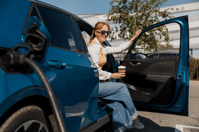 Sieviete sēž elektroauto ar kafijas krūzi un gaida, kamēr viņas elektroauto uzlādēsies.