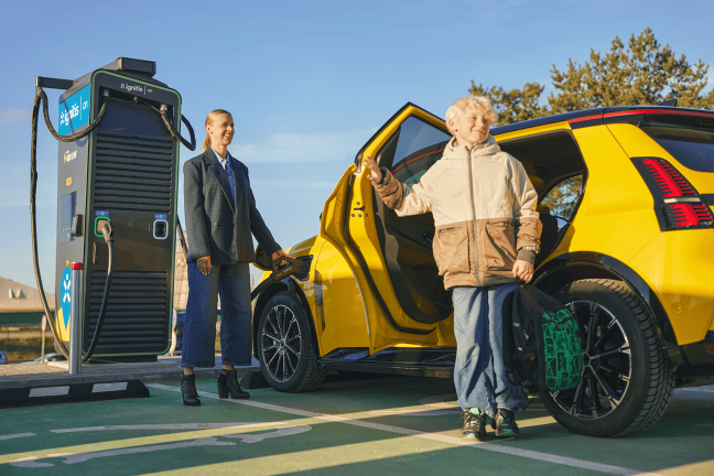Sieviete uzlādē savu dzelteno elektroauto Ignitis ON īpaši ātrās uzlādes stacijā, pie elektroauto stāv bērns ar atvērtām durvīm.