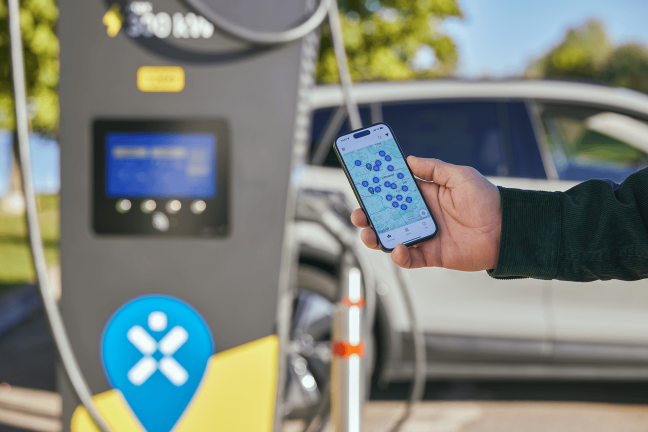 Vīrietis stāv blakus Ignitis ON elektroauto uzlādes stacijai un rokā tur tālruni, kurā ir atvērta Ignitis ON mobilā lietotne.